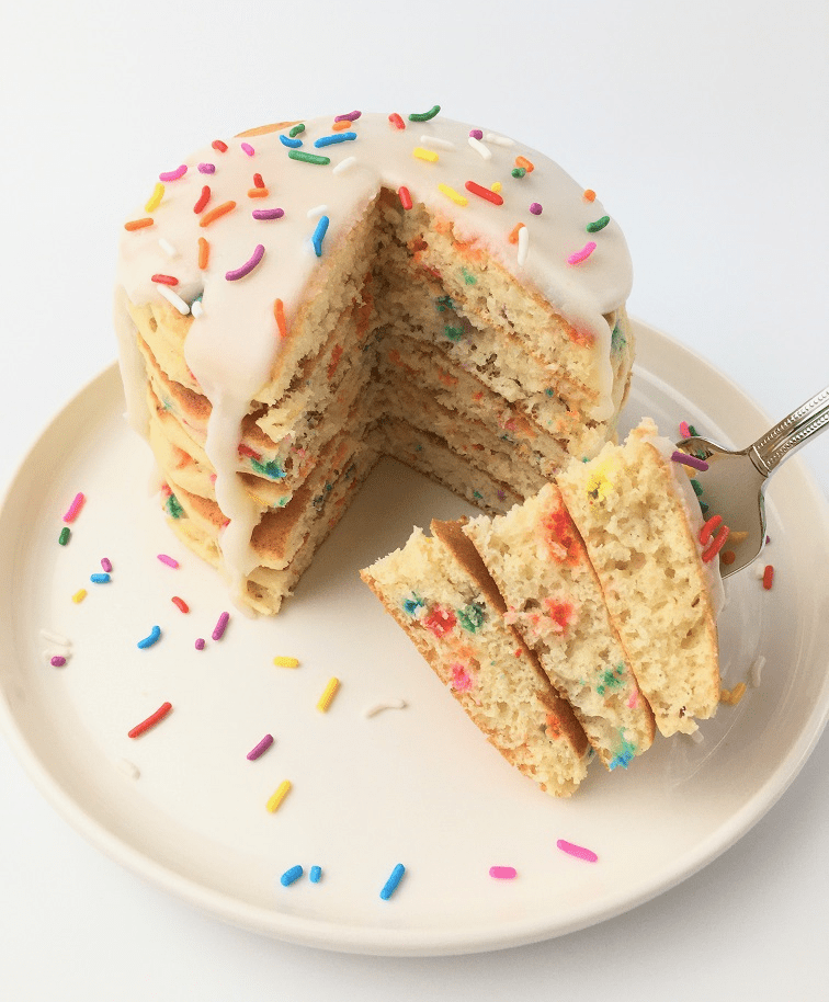 Stack of finished birthday funfetti pancakes topped with white icing and extra rainbow sprinkles with a cut wedge on a fork