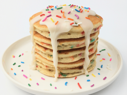 Stack of finished birthday funfetti pancakes topped with white icing and extra rainbow sprinkles