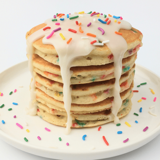 Stack of finished birthday funfetti pancakes topped with white icing and extra rainbow sprinkles