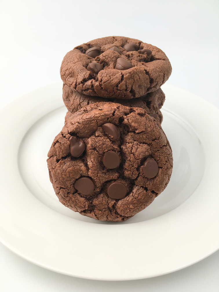 One double chocolate chip cookie leaning against a stack of cookies on a white plate