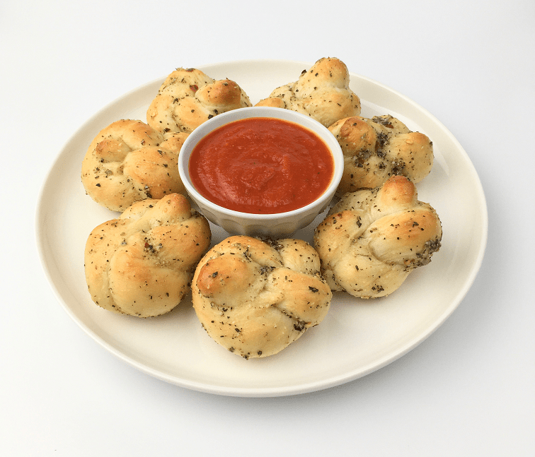 Baked garlic knots arranged on a white plate around a small bowl of pizza sauce