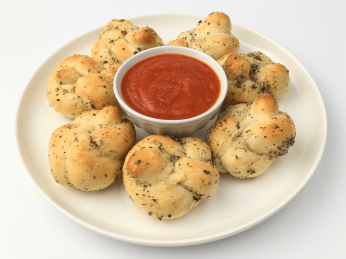 Baked garlic knots arranged on a white plate around a small bowl of pizza sauce