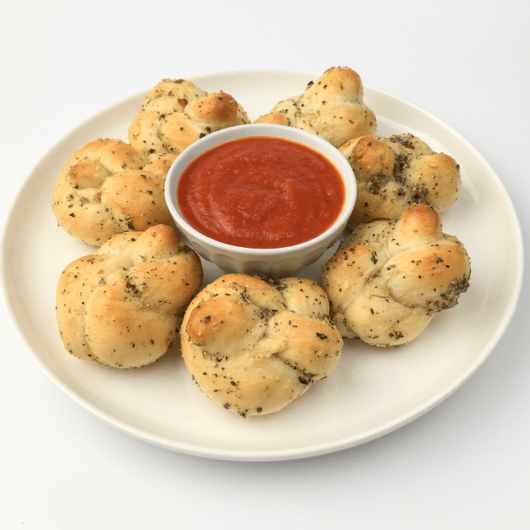 Baked garlic knots arranged on a white plate around a small bowl of pizza sauce