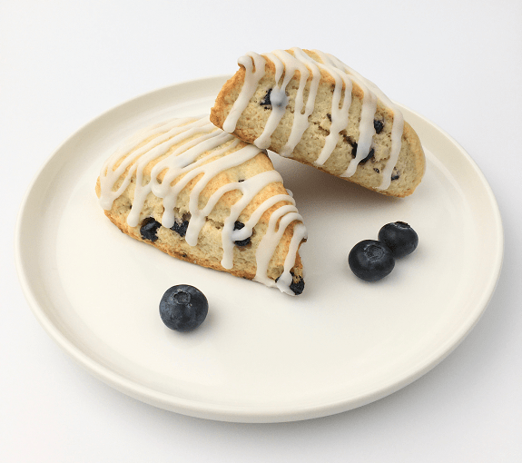 Two glazed lemon blueberry scones on a white plate garnished with fresh blueberries