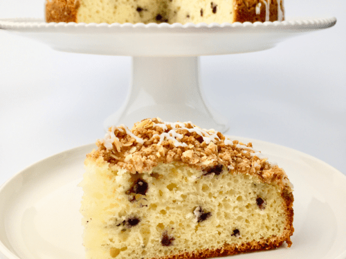 Slice of blueberry streusel muffin cake on a white plate in front of the cake stand