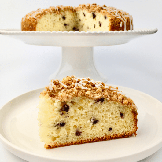 Slice of blueberry streusel muffin cake on a white plate in front of the cake stand