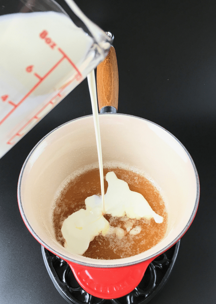 Pouring heavy cream from a measuring cup into the caramelized sugar in a saucepan