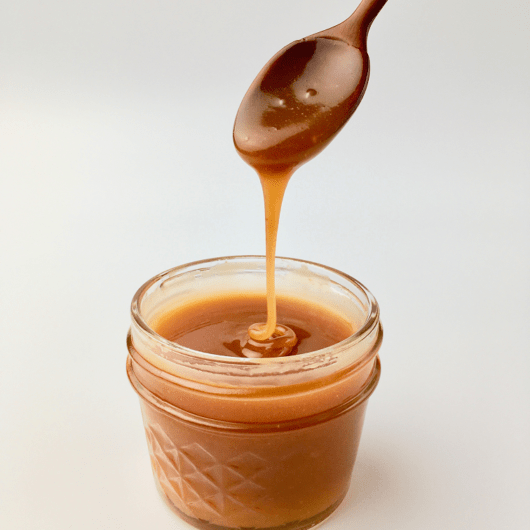 Homemade caramel sauce dripping off a wooden spoon into a glass jar