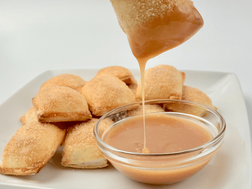 Apple pie bites on a plate with one dipped in caramel sauce.