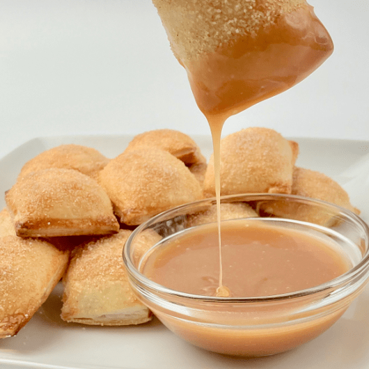 Apple pie bites on a plate with one dipped in caramel sauce.