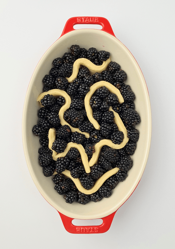 Fresh blackberries arranged around strips of cobbler dough in a red casserole dish