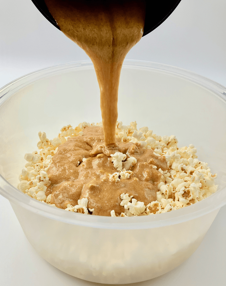 Pouring maple spice caramel over popcorn in plastic bowl.