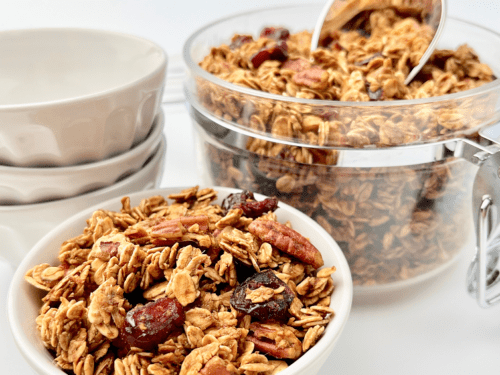 Gingerbread granola in a white bowl and glass canister with a scoop.