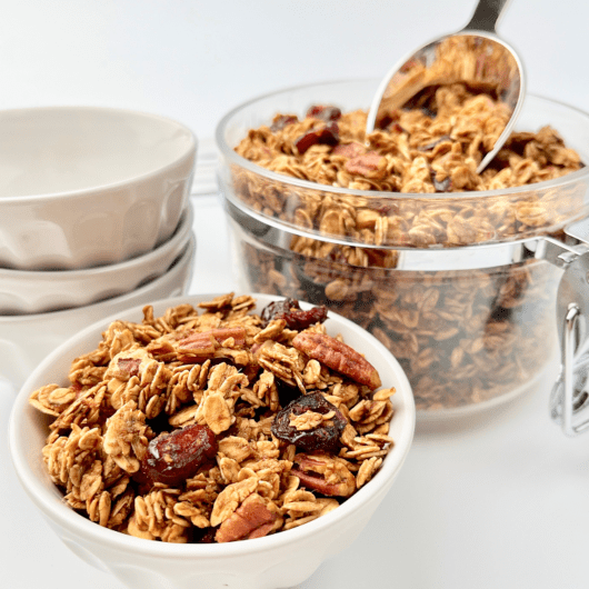 Gingerbread granola in a white bowl and glass canister with a scoop.